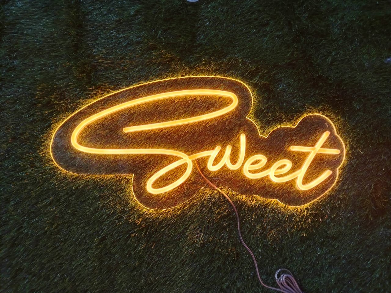 Neon sign displaying the word "Sweet" in glowing pink or pastel light with a playful, soft font.